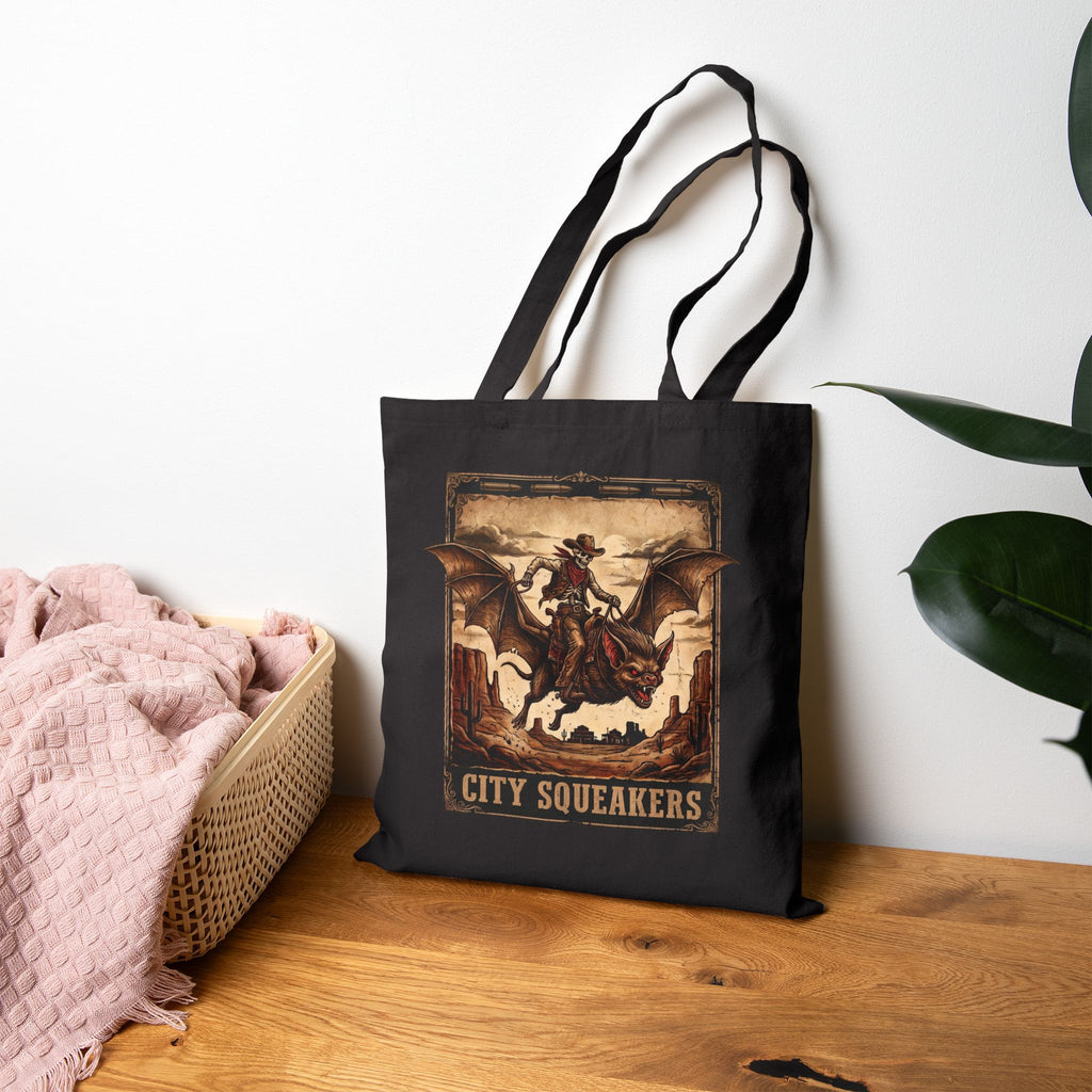 A black tote bag with a vintage-style 'City Squeakers' graphic print, displayed on a wooden table next to a pink blanket and a green plant against a white wall.