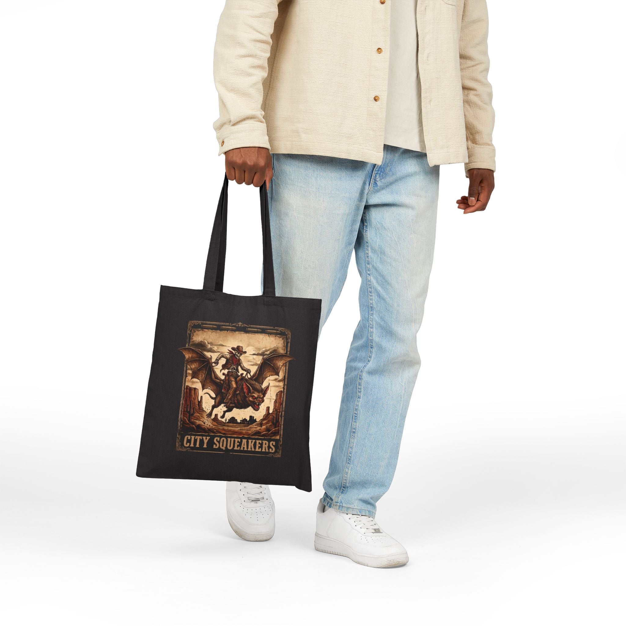 Man holding black City Squeakers tote bag front view — vintage skeleton cowboy bat rider graphic, heavy cotton shoulder bag for everyday carry