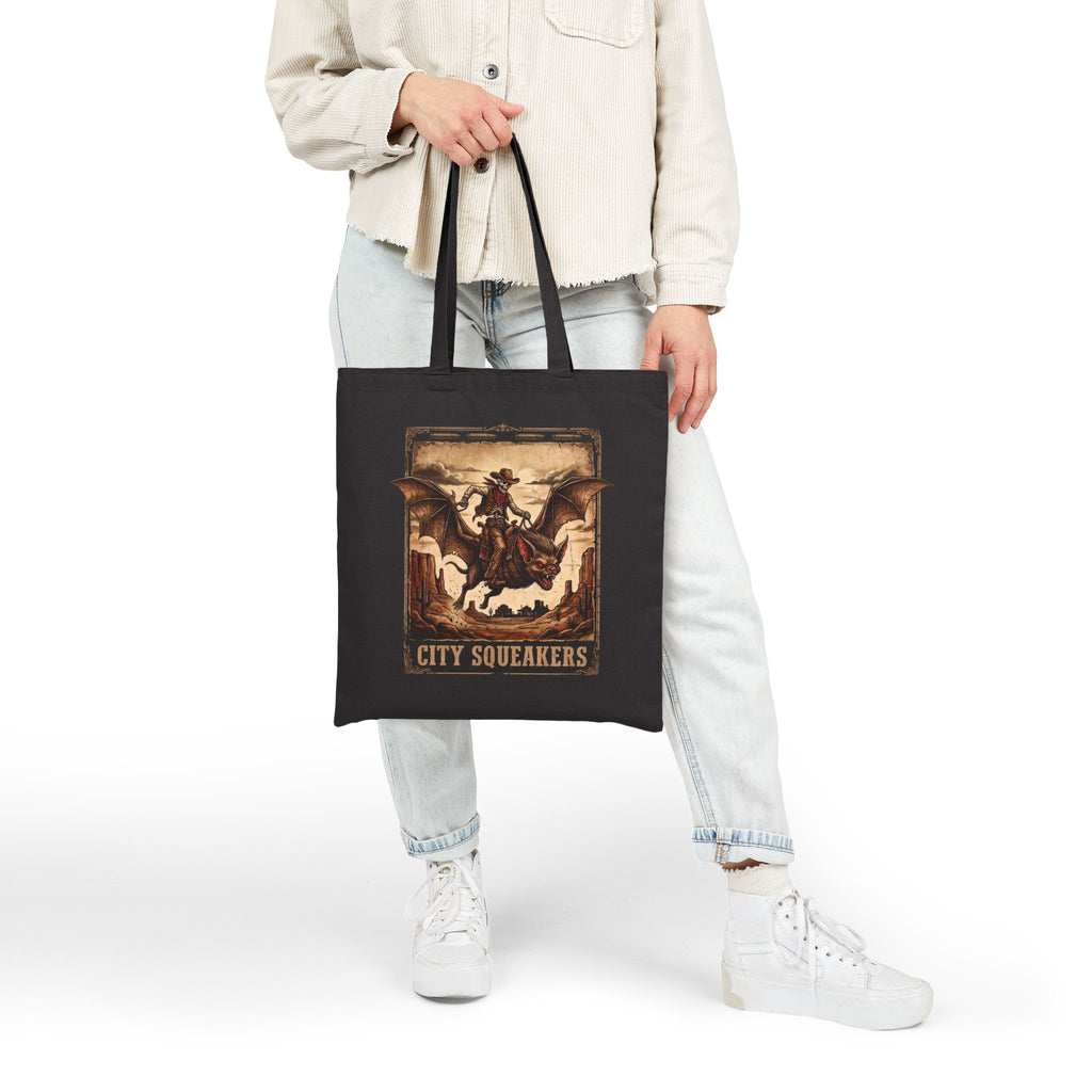 Woman holding black City Squeakers tote bag front view — vintage skeleton cowboy bat rider graphic, heavy cotton shoulder bag for everyday carry