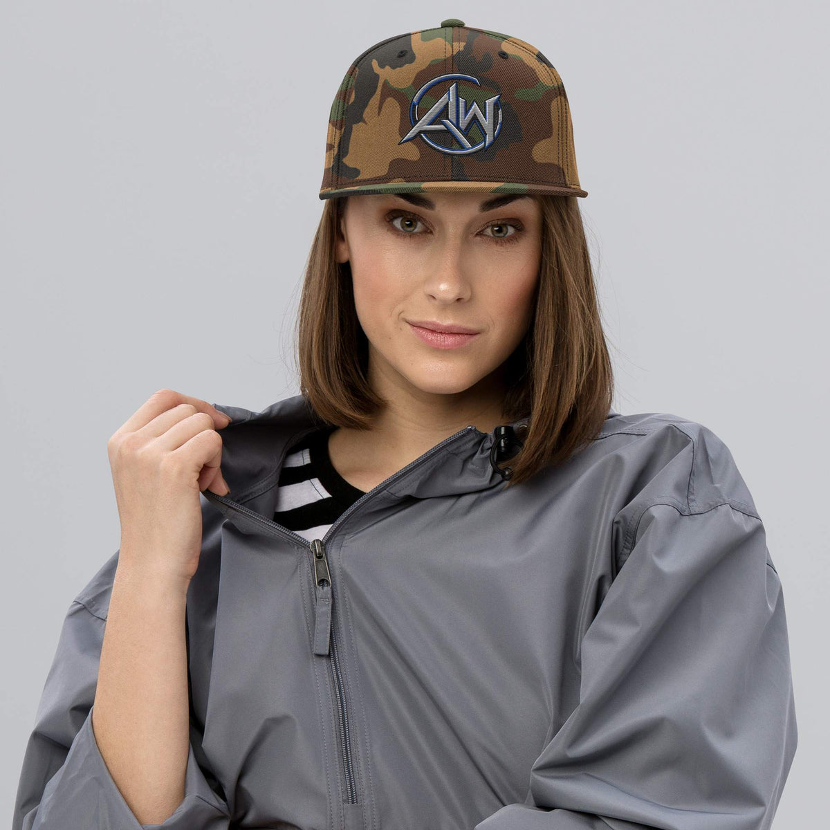 Woman wearing a gray raincoat and camouflage cap with an AW logo on a gray background