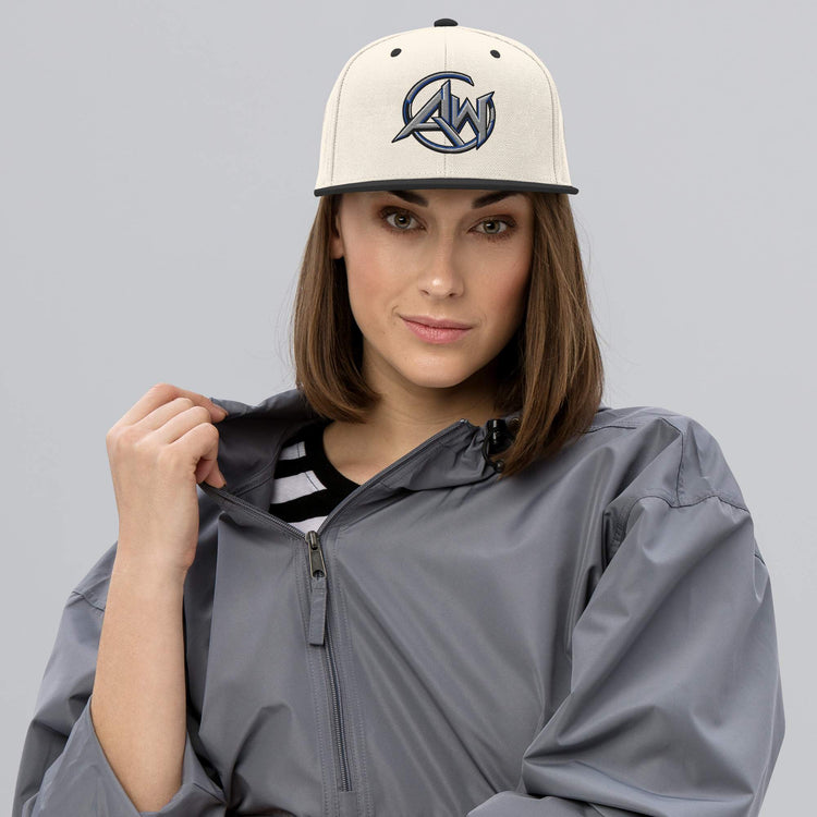 A woman wearing a cream snapback cap with a dark blue 'AW' logo and a gray zip-up jacket, photographed against a solid gray studio background.
