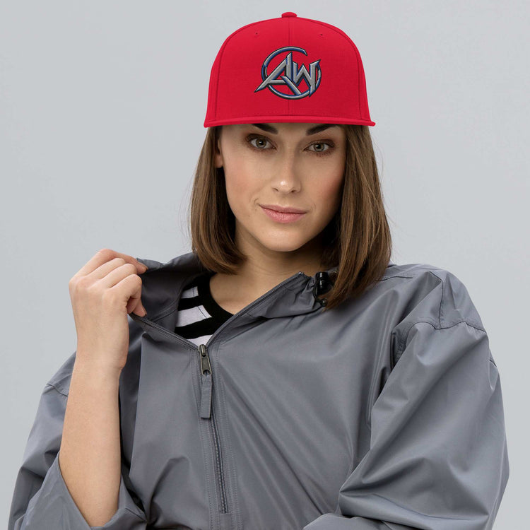 A woman wearing a bright red snapback cap with a stylized 'AW' logo in black and gray, paired with a gray zip-up jacket over a striped shirt, set against a solid gray studio background.