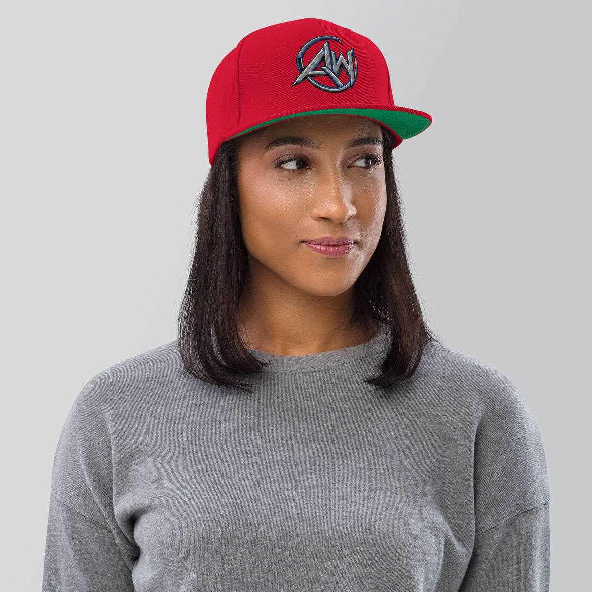 A woman wearing a red baseball cap with a green brim and a circular silver  AW logo on the front, paired with a gray crewneck sweatshirt, set against a solid light gray background.