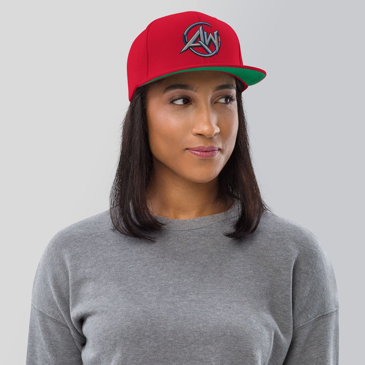 A woman wearing a red baseball cap with a green brim and a circular silver  AW logo on the front, paired with a gray crewneck sweatshirt, set against a solid light gray background.
