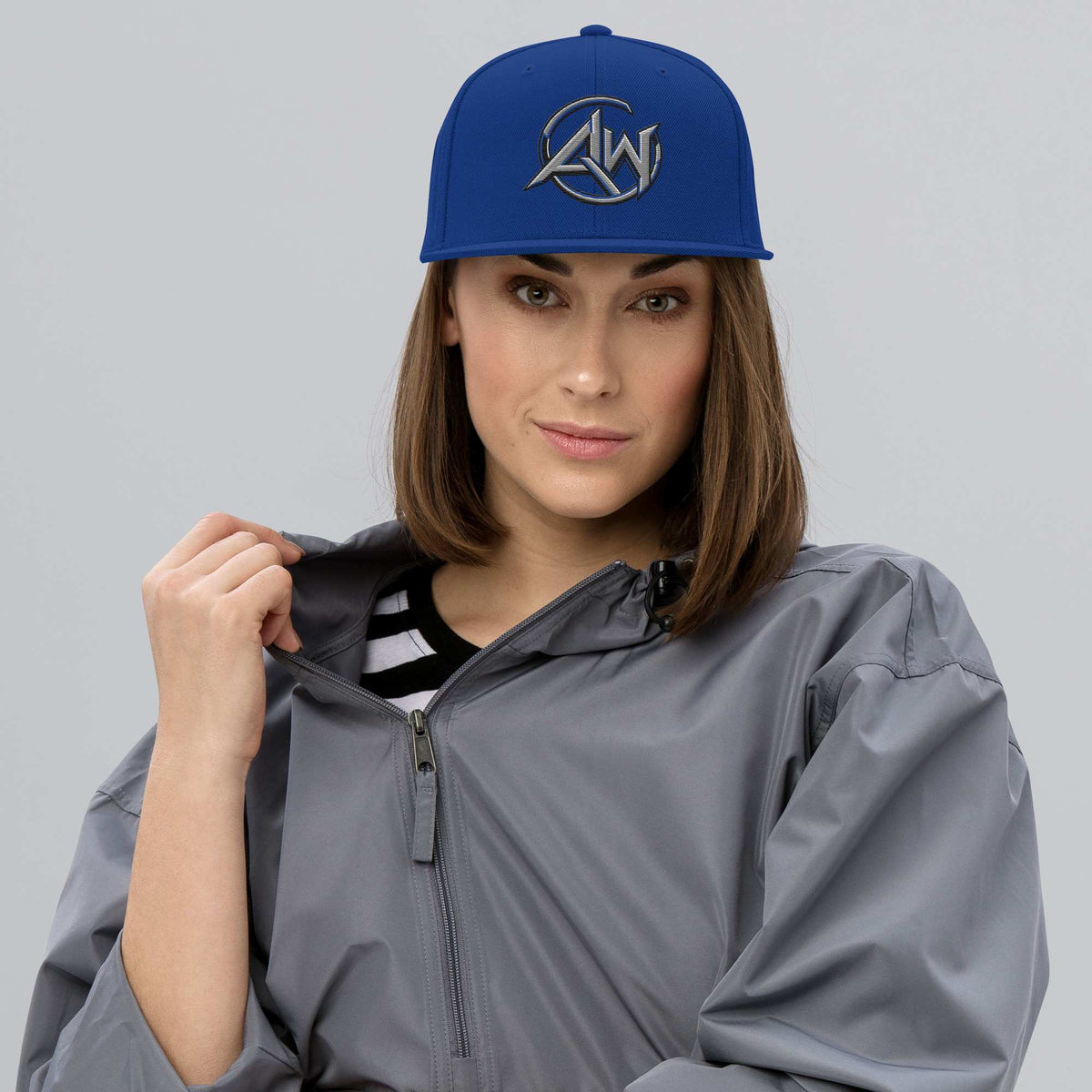 Woman wearing a blue cap with An AW logo and a gray raincoat on a light gray background
