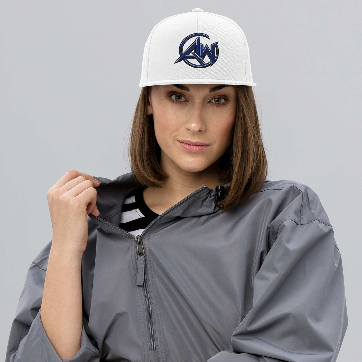 A woman wearing a white snapback cap with a navy blue AW logo and a gray zip-up jacket, photographed against a solid gray studio background.