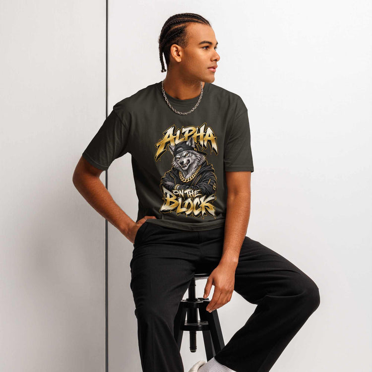 A man wearing a dark charcoal gray graphic t-shirt with a gold and black 'Alpha on the  Block' wolf design, paired with black pants, sitting on a black stool against a light gray wall.