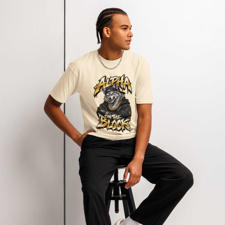 A man wearing a cream-colored graphic t-shirt with a wolf and the text 'Alpha on the Block' man sits on a black stool against a light gray wall.