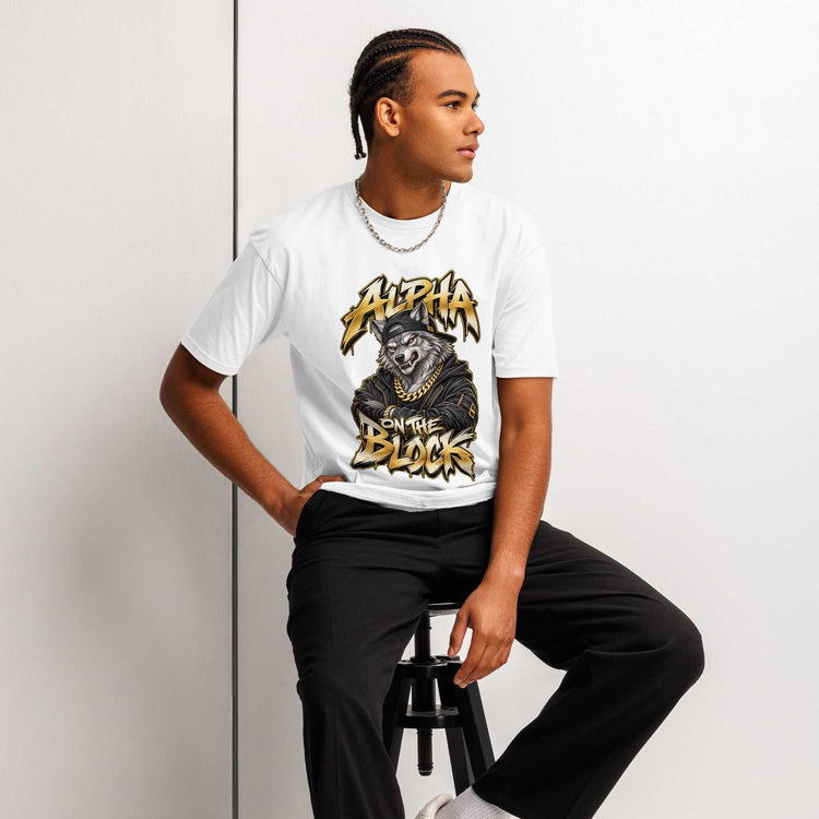 A man wearing a white graphic t-shirt with a wolf illustration and the text ' ALPHA ON THE BLOCK' in gold and black, paired with black pants, sitting on a stool against a white wall.