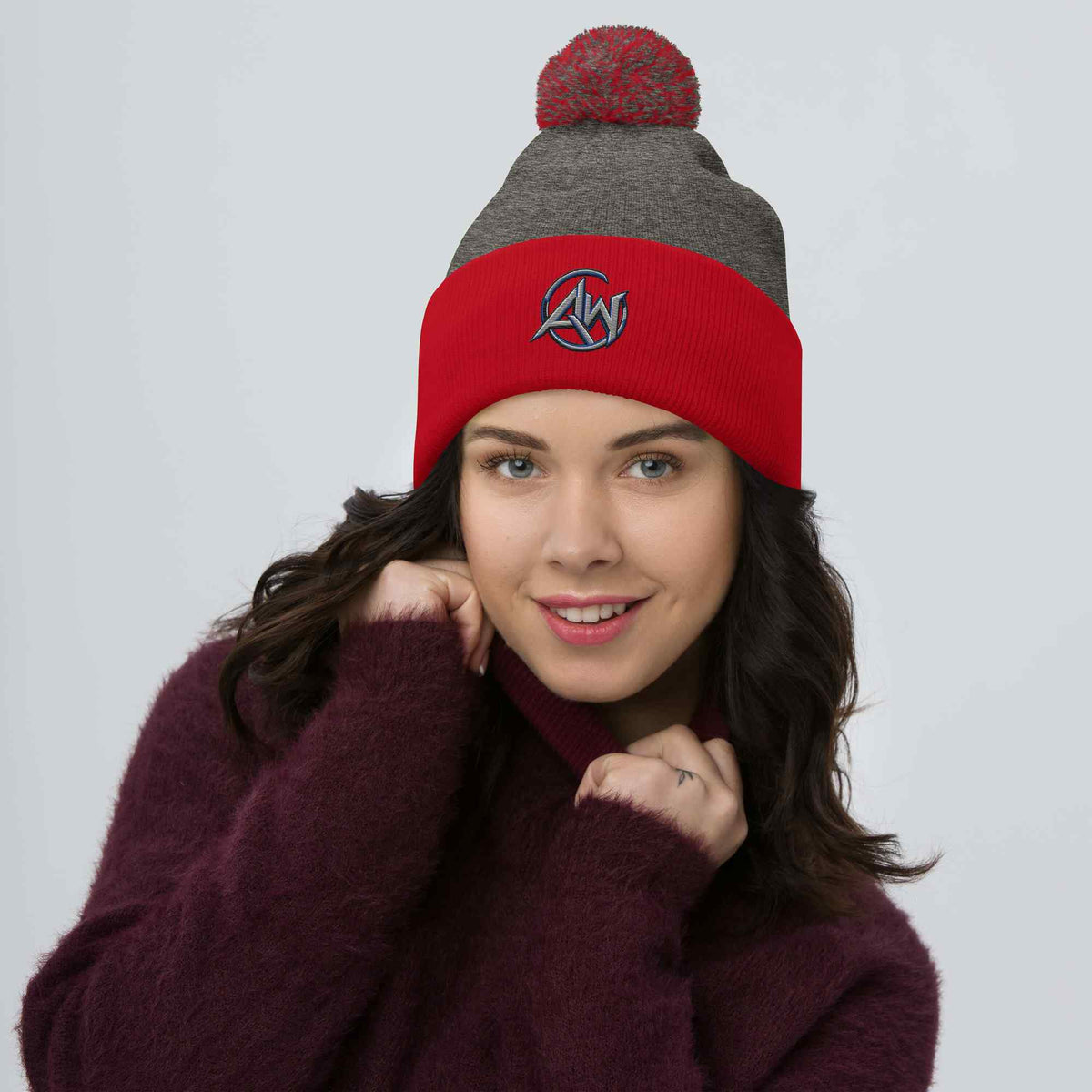 Woman wearing a red and gray beanie pom pom with an AW logo on a plain background