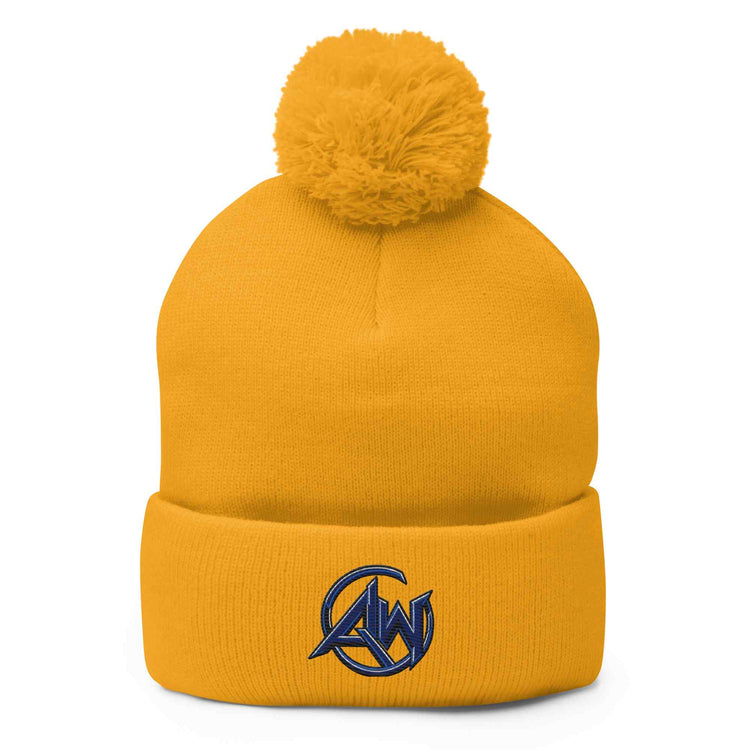 A mustard yellow knit pom-pom beanie with a folded cuff and a navy blue embroidered circular AW logo on the front, shown against a plain white background.