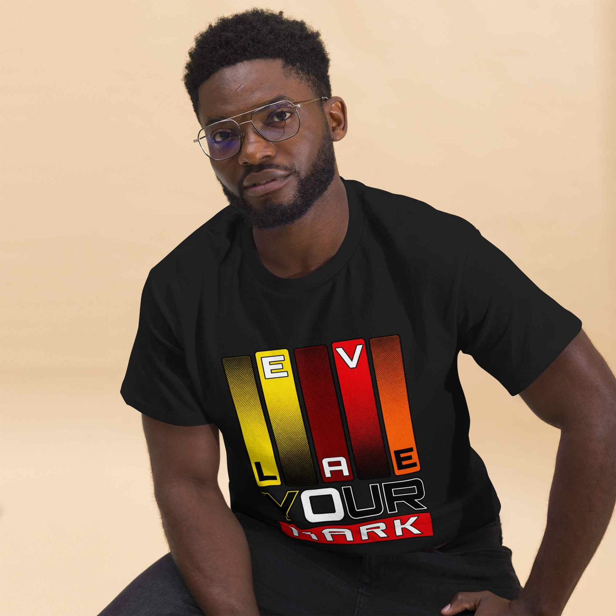A man wearing a black t-shirt with a colorful graphic print that reads 'LEAVE YOUR MARK' in yellow, red, orange, and white, posed against a solid beige background.
