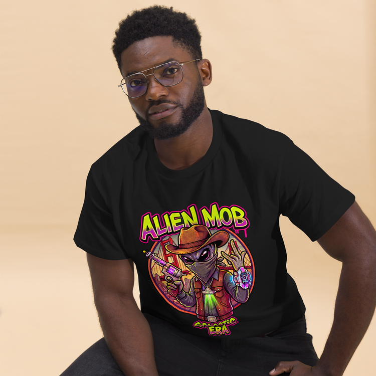 A man wearing a black graphic t-shirt with a western  alien cowboy design and the word 'Alien Mob Galactic ERA ' printed on the front, posed against a solid beige background.