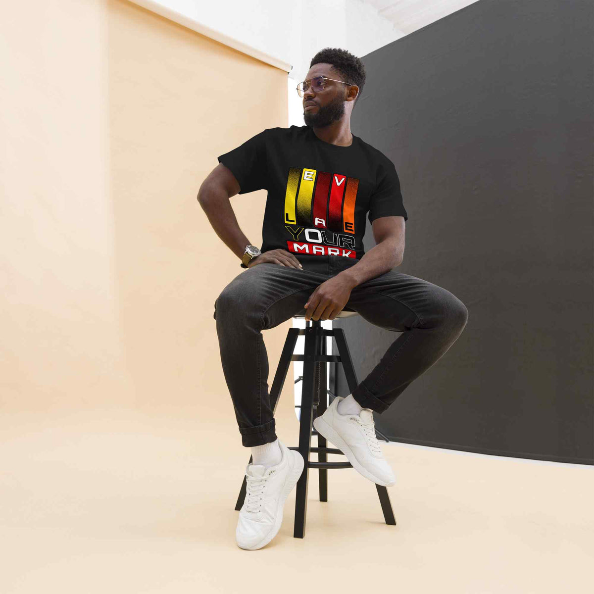Man sitting on a stool wearing a black t-shirt with a text Leave your mark  design graphic, black pants, and white sneakers.