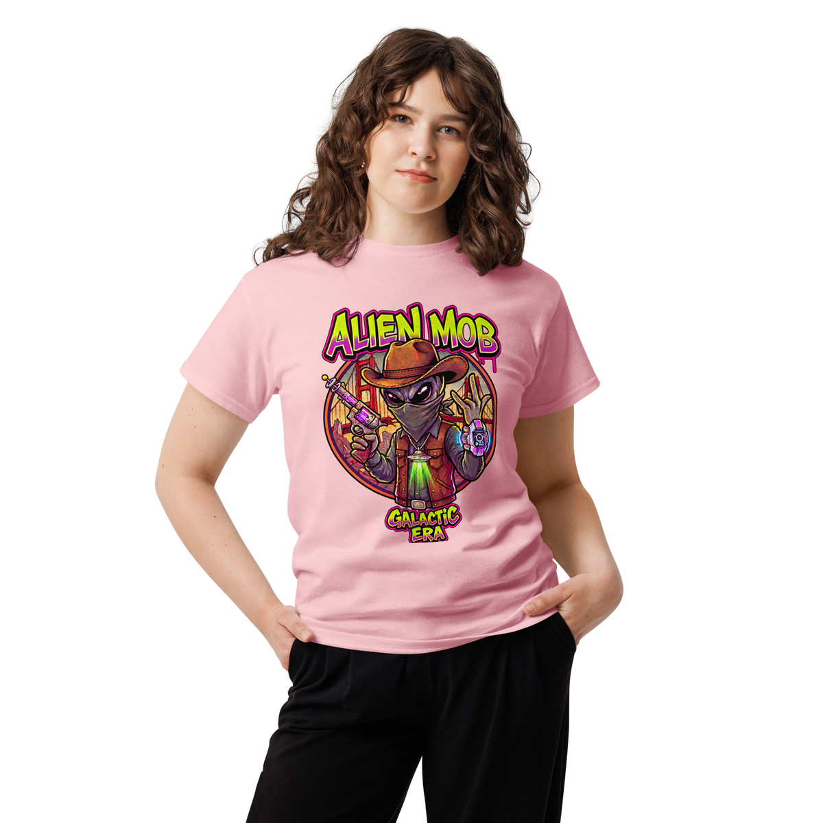 A young woman with wavy brown hair wearing a light pink graphic  Alien mob alien cowboy outlaw t-shirt and black pants, standing against a plain white background.