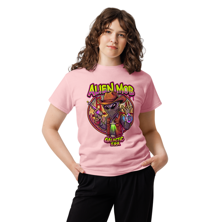 A young woman with wavy brown hair wearing a light pink graphic  Alien mob alien cowboy outlaw t-shirt and black pants, standing against a plain white background.