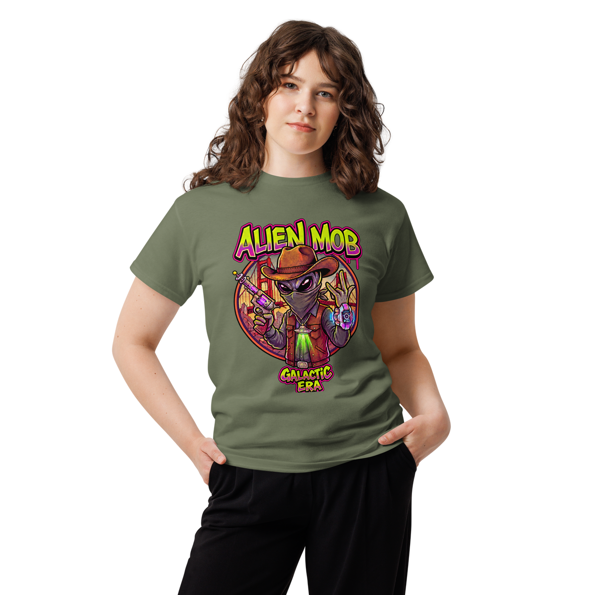 A woman modeling an olive green graphic t-shirt with a colorful 'Alien Mob Galactic Era' design on the front, paired with black pants, against a plain white background.
