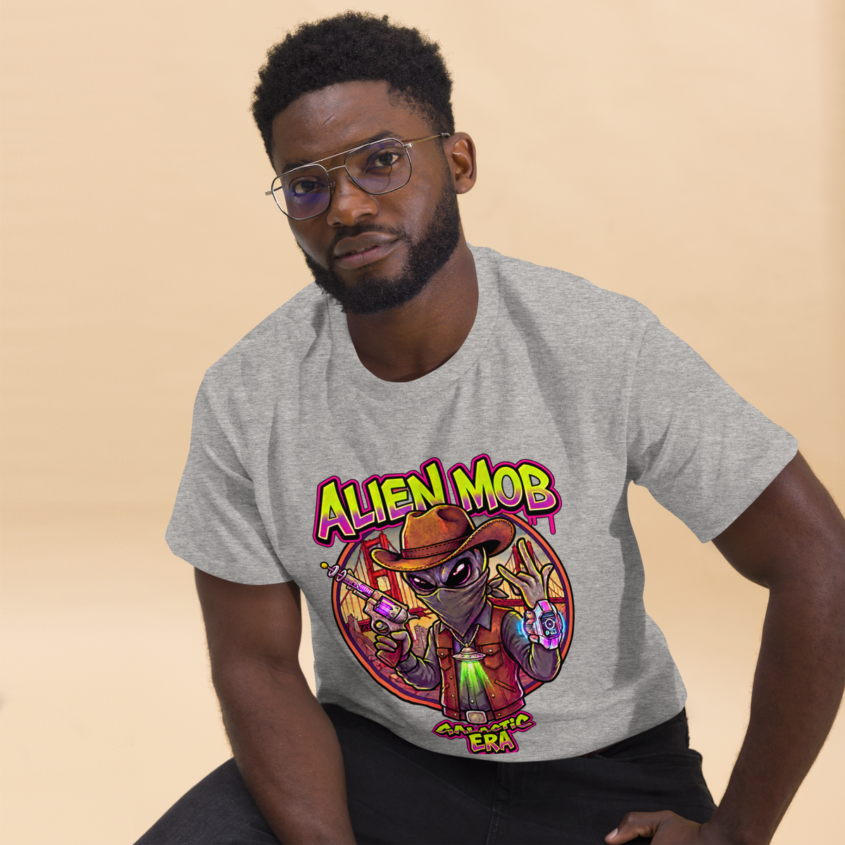 A man wearing a light gray heather t-shirt with a colorful cartoon alien graphic that reads 'ALIEN MOB' GALACTIC ERA'.