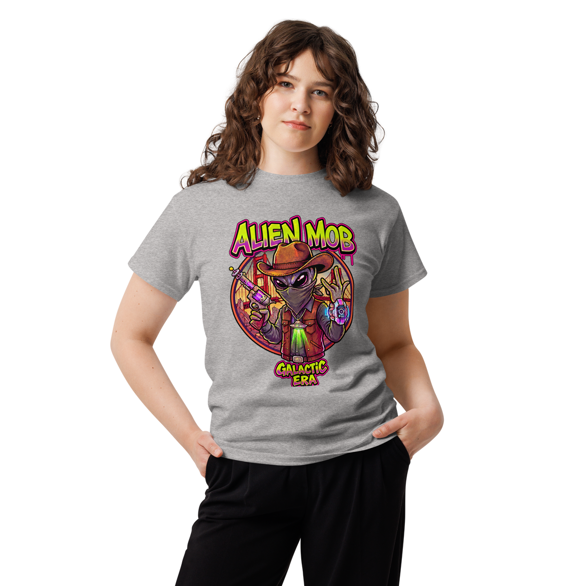 A young woman with wavy brown hair wearing a heather gray graphic t-shirt and black pants, standing against a plain white background. The t-shirt features a colorful graphic of a cartoon alien wearing a cowboy hat, with the text 'ALIEN MOB' and 'GALACTIC ERA'.