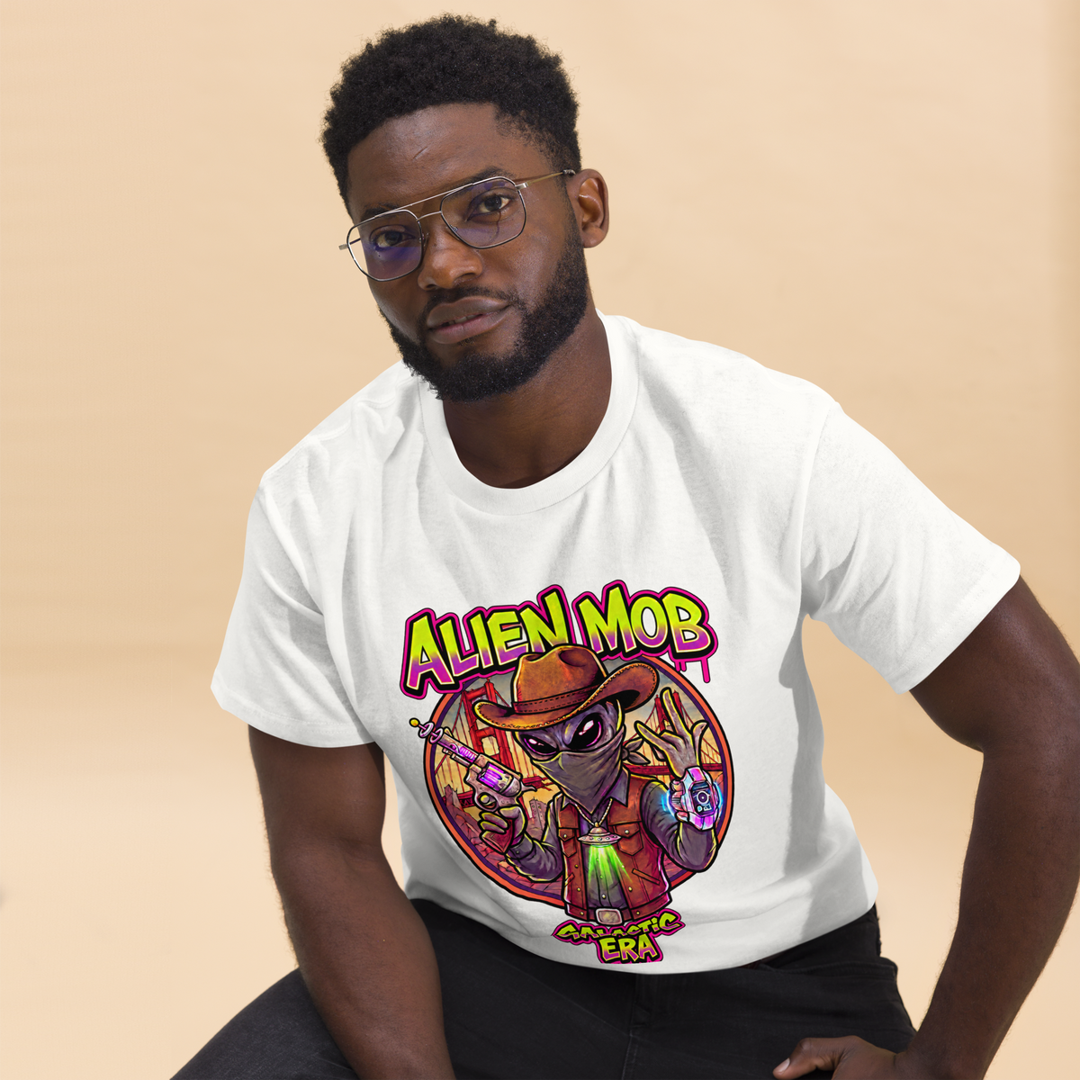 A man wearing a white graphic t-shirt with a colorful cartoon alien design and the text 'ALIEN MOB ERA against a solid beige background.
