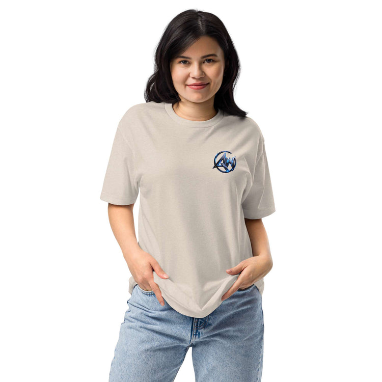 A woman modeling a light beige crewneck t-shirt with a small blue circular  logo AW text graphic on the left chest, paired with light blue jeans, against a plain white background.