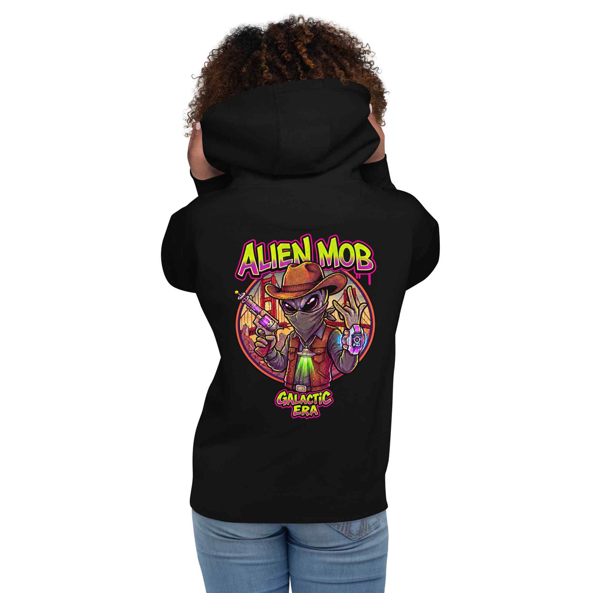 woman wearing a black hoodie with alien mob cowboy outlaw  on the back of the hoodie paired with blue jeans on a white background back view