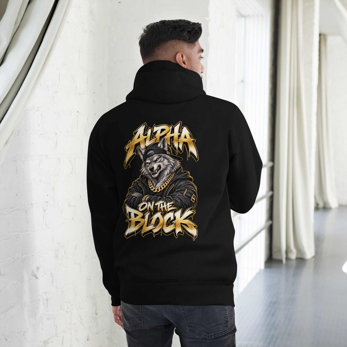 A man viewed from behind wearing a black hoodie with a large graphic print on the back. The print features a wolf wearing a gold chain and the text 'ALPHA ON THE BLOCK' in a stylized gold font.