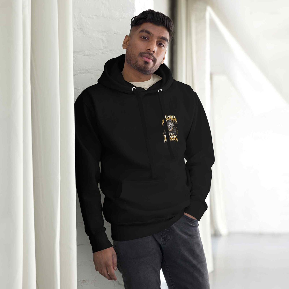 A man wearing a black pullover hoodie with a gold  Alpha On The Block graphic wolf print on the left chest, standing in a bright, modern room with white walls and curtains.