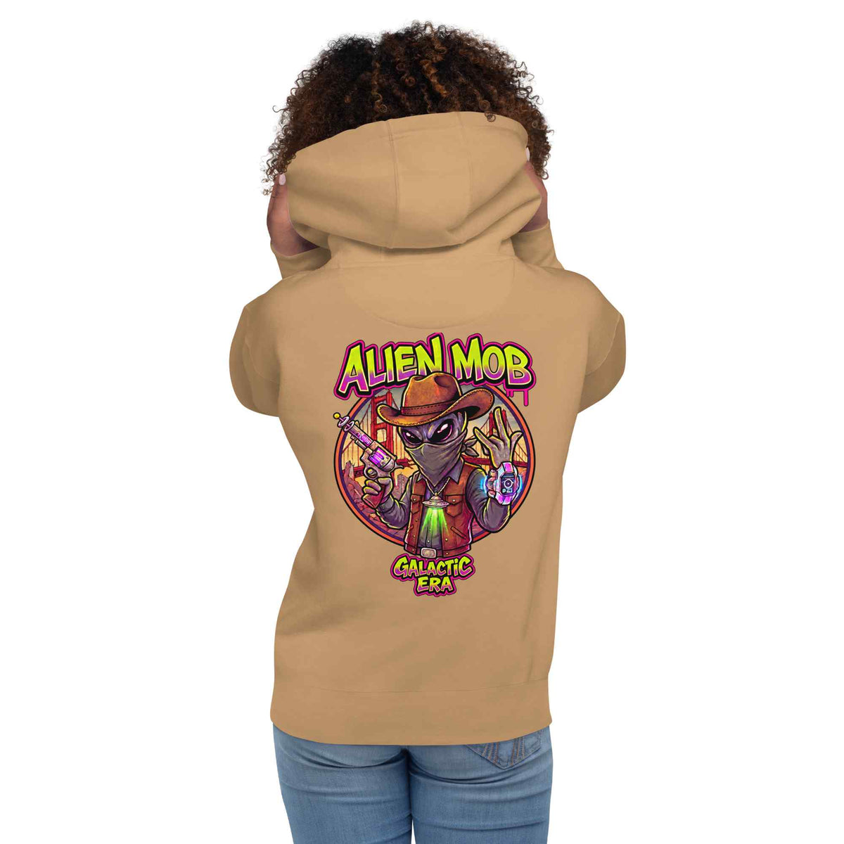 woman wearing a khaki hoodie with alien mob cowboy outlaw  on the back of the hoodie paired with blue jeans on a white background back view