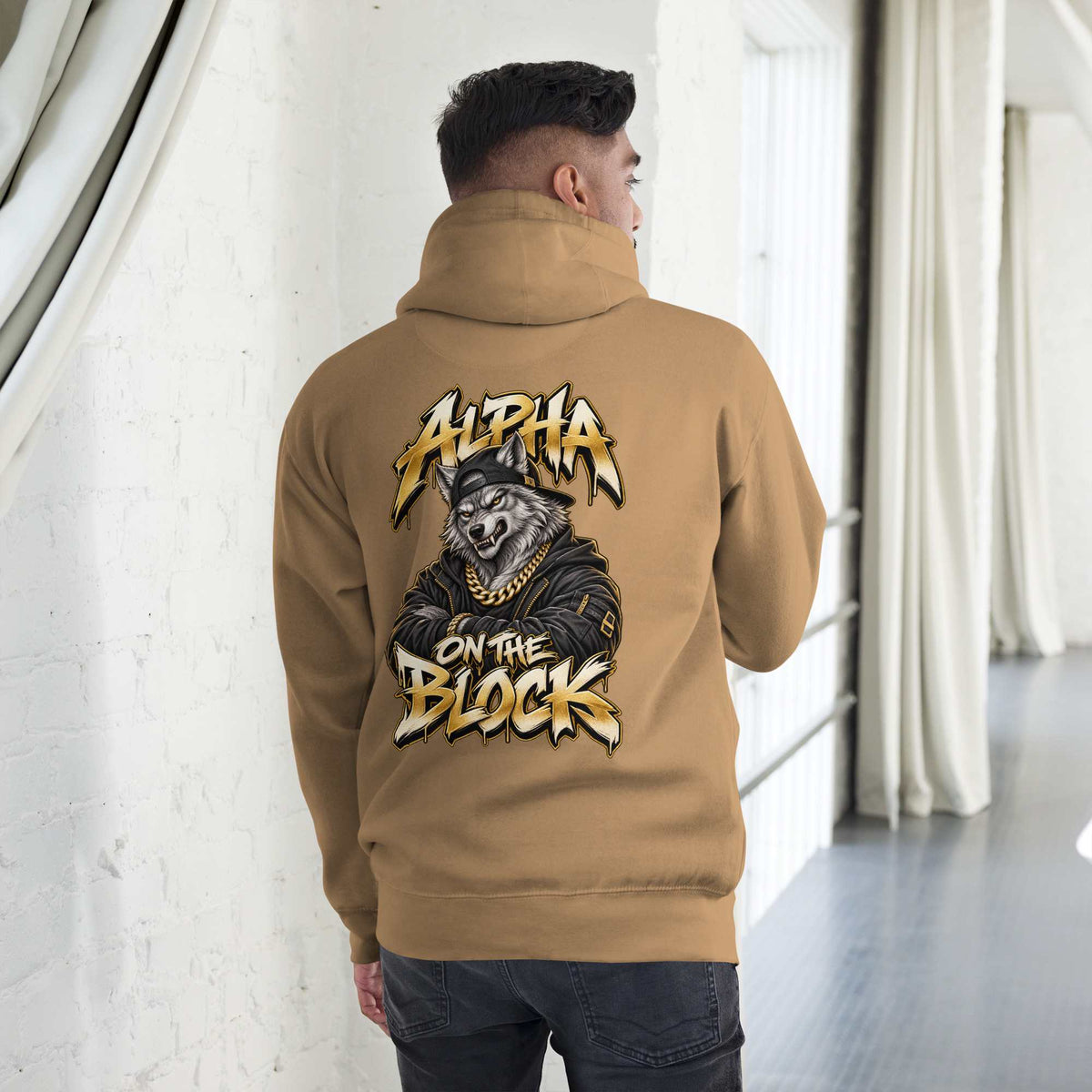 Person wearing a tan hoodie with a wolf design on the back, standing in a room with white walls and a window.