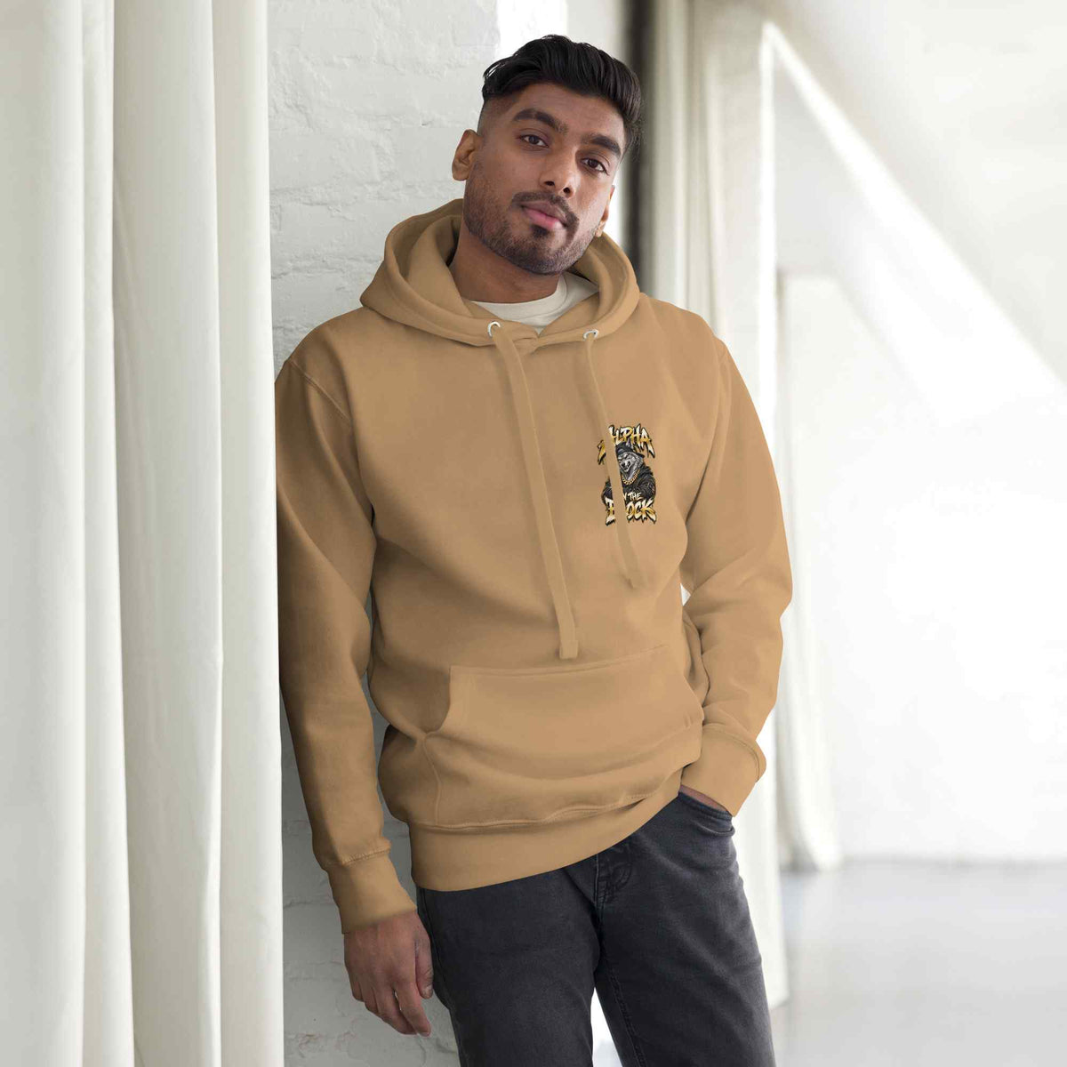 A man wearing a tan pullover hoodie with a black graphic of a wolf, and the text Alpha On The Block leaning against a white wall in a bright indoor setting.