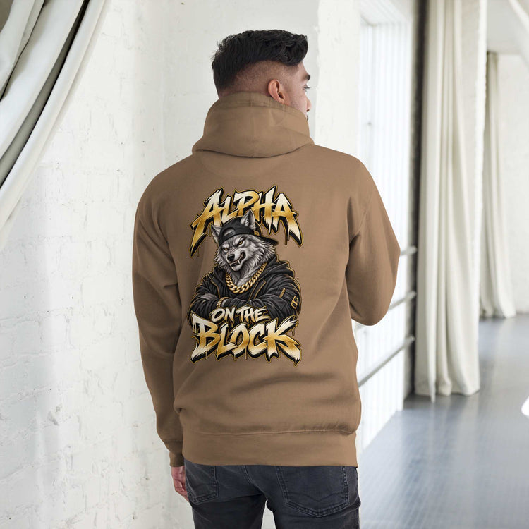 A man viewed from behind wearing a tan hoodie with a large graphic on the back featuring a wolf in a cap and the text 'ALPHA ON THE BLOCK' in stylized yellow and white lettering. The setting is a bright, modern indoor space with white walls and curtains.