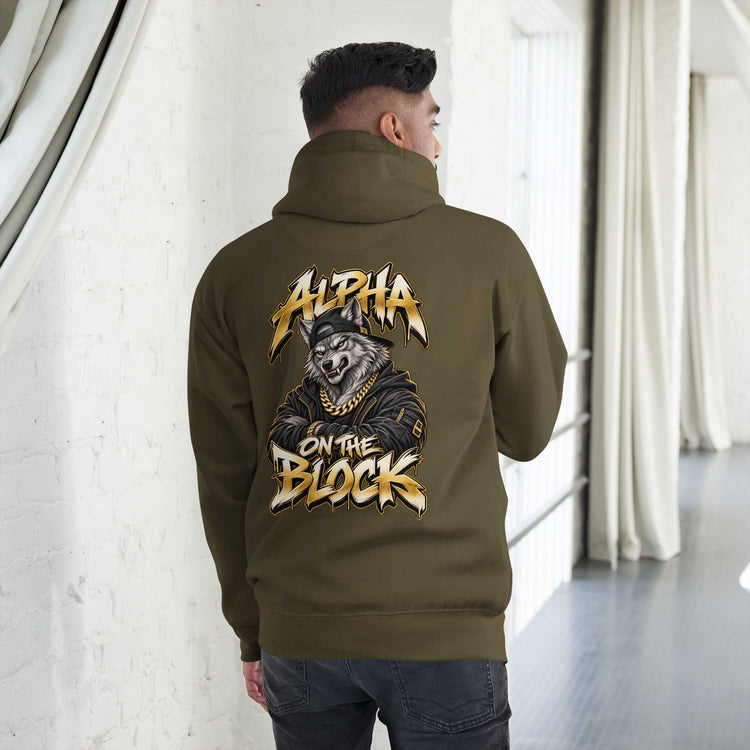 A man viewed from behind wearing an olive green hoodie with a large graphic on the back featuring a wolf in a leather jacket and the text 'ALPHA ON THE BLOCK' in gold and white. He is standing in a bright indoor hallway with white walls and curtains.