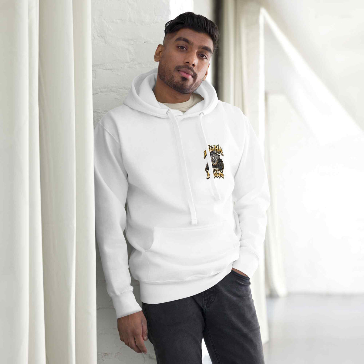 A man wearing a white pullover hoodie with a small wolf  graphic print on the left chest, posed in a bright, modern room with white curtains and a white brick wall.