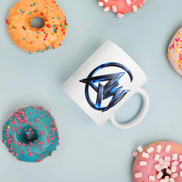 A white ceramic coffee mug with a metallic blue and black stylized 'AW' logo, presented in a flat-lay arrangement on a light blue background with colorful donuts.