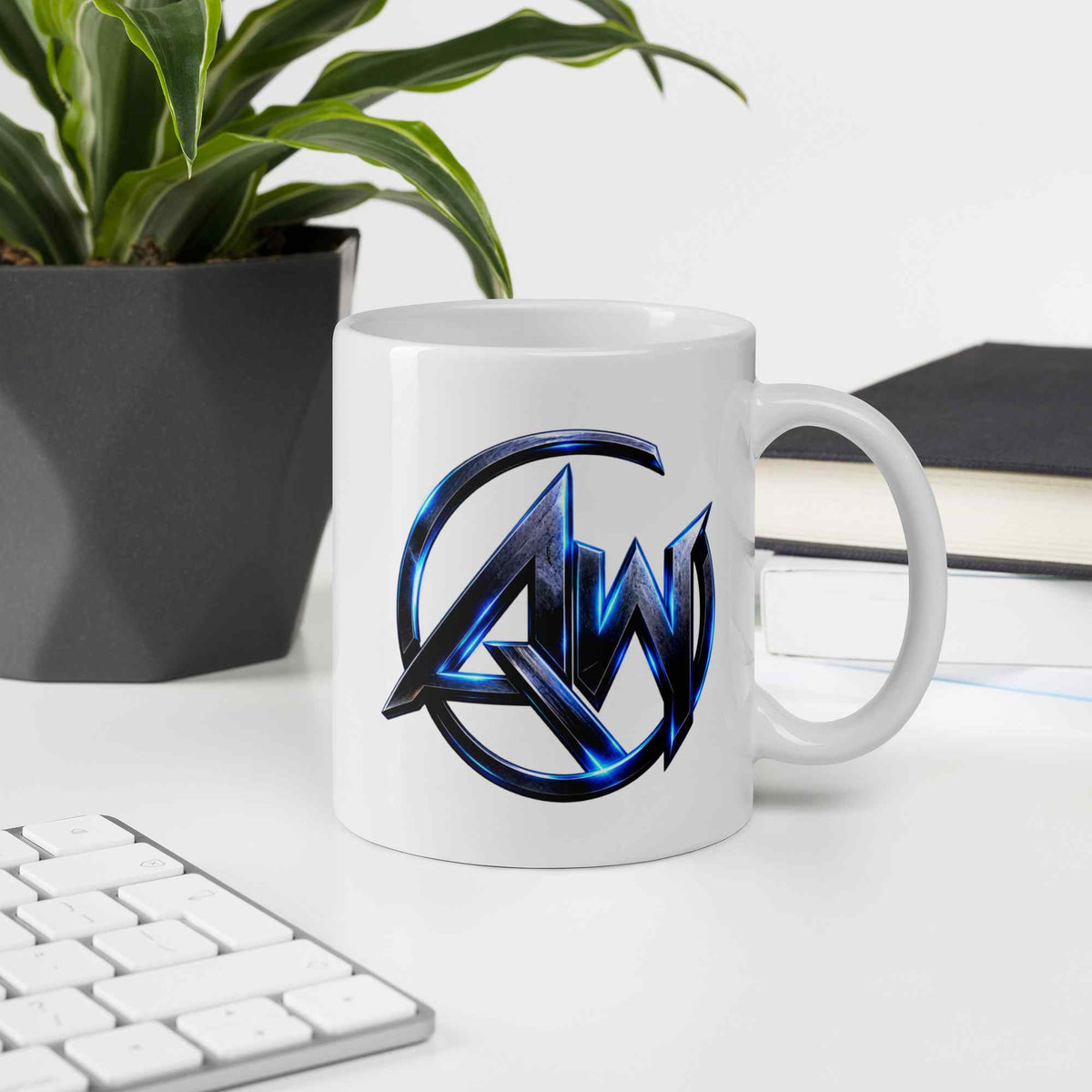 A white ceramic coffee mug with a metallic blue and black AW logo, displayed on a white desk next to a keyboard, a potted plant, and a notebook.