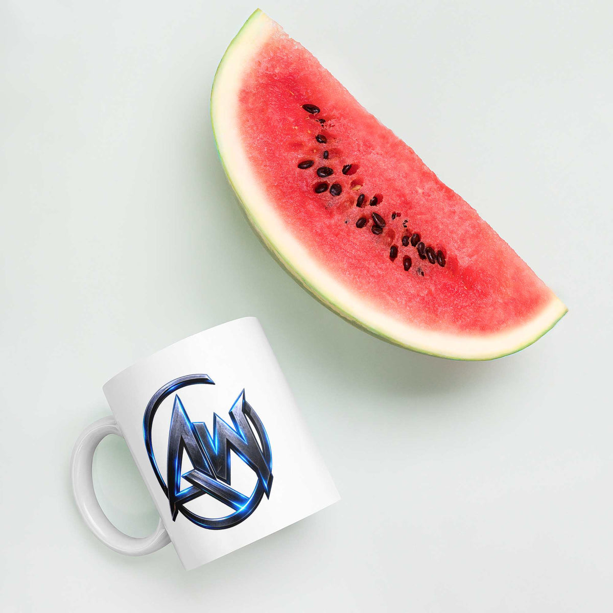A white ceramic mug with a blue and black AW-style logo, shown next to a slice of watermelon on a light gray background.