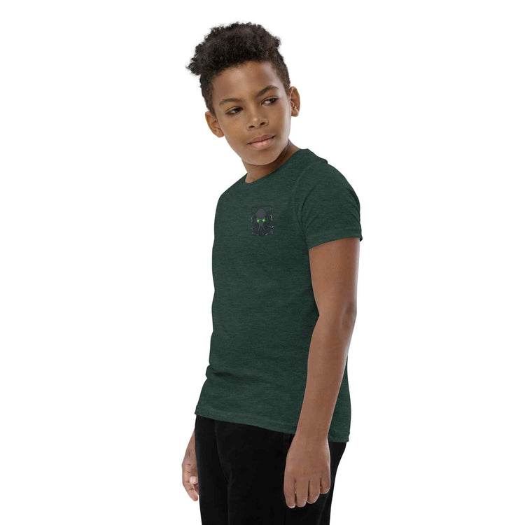 A child model wearing a dark green heathered t-shirt with small kraken graphics on the left chest, paired with black pants, set against a plain white background.