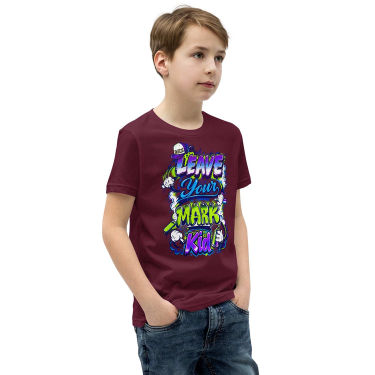 A young boy wearing a maroon t-shirt with a colorful graphic that reads 'LEAVE YOUR MARK KID' in stylized lettering, paired with blue jeans, set against a plain white background.