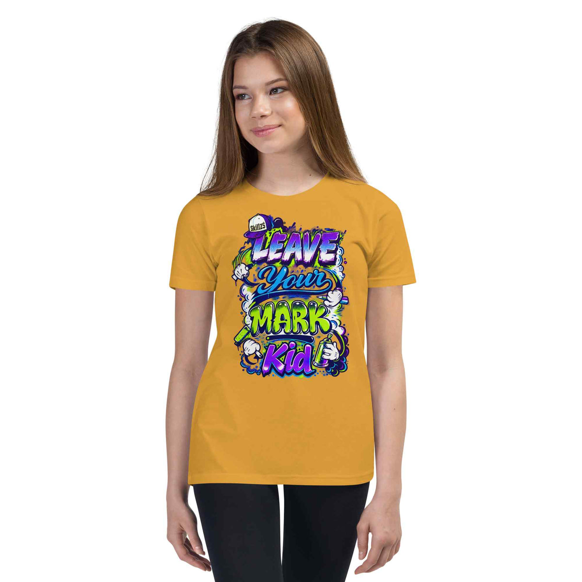 A young woman modeling a mustard yellow t-shirt with a colorful graphic that reads 'LEAVE YOUR MARK Kid' in purple, green, and blue stylized lettering, set against a plain white background.