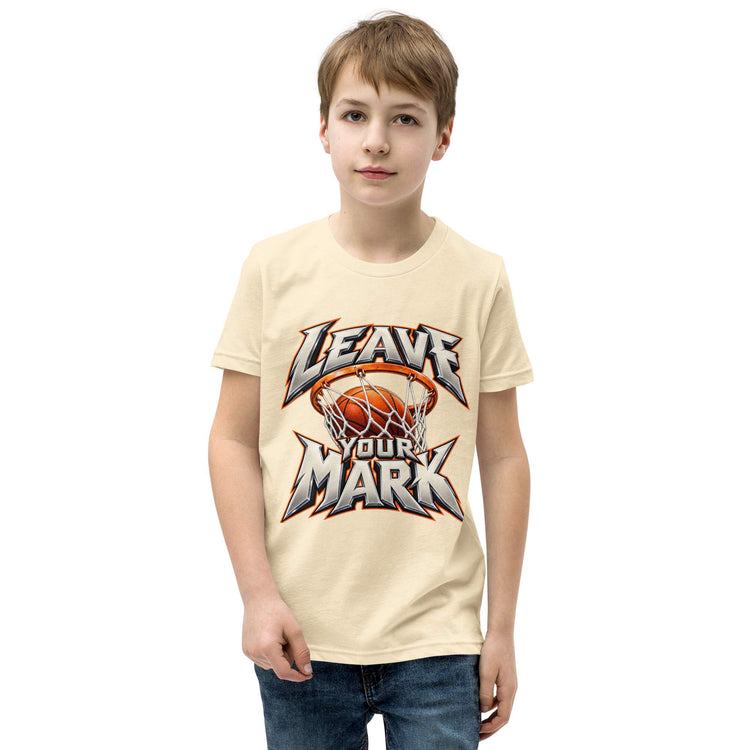 A young boy wearing a light beige graphic t-shirt with a basketball-themed 'LEAVE YOUR MARK' design, paired with blue jeans, set against a plain white background.