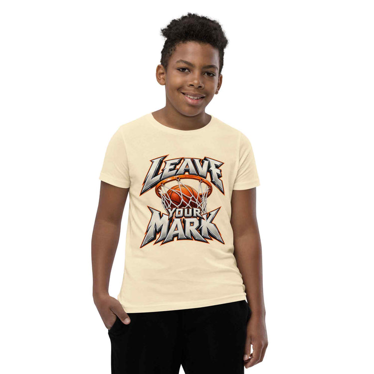 A young boy wearing a cream-colored t-shirt with a basketball-themed graphic that reads 'LEAVE YOUR MARK'. The background is solid white.