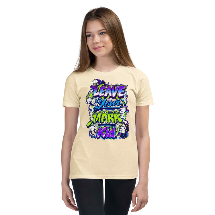 A young woman modeling a light beige t-shirt with a colorful graphic that reads 'LEAVE YOUR MARK Kid' in stylized purple, blue, and green lettering. She is wearing black pants against a solid white background.