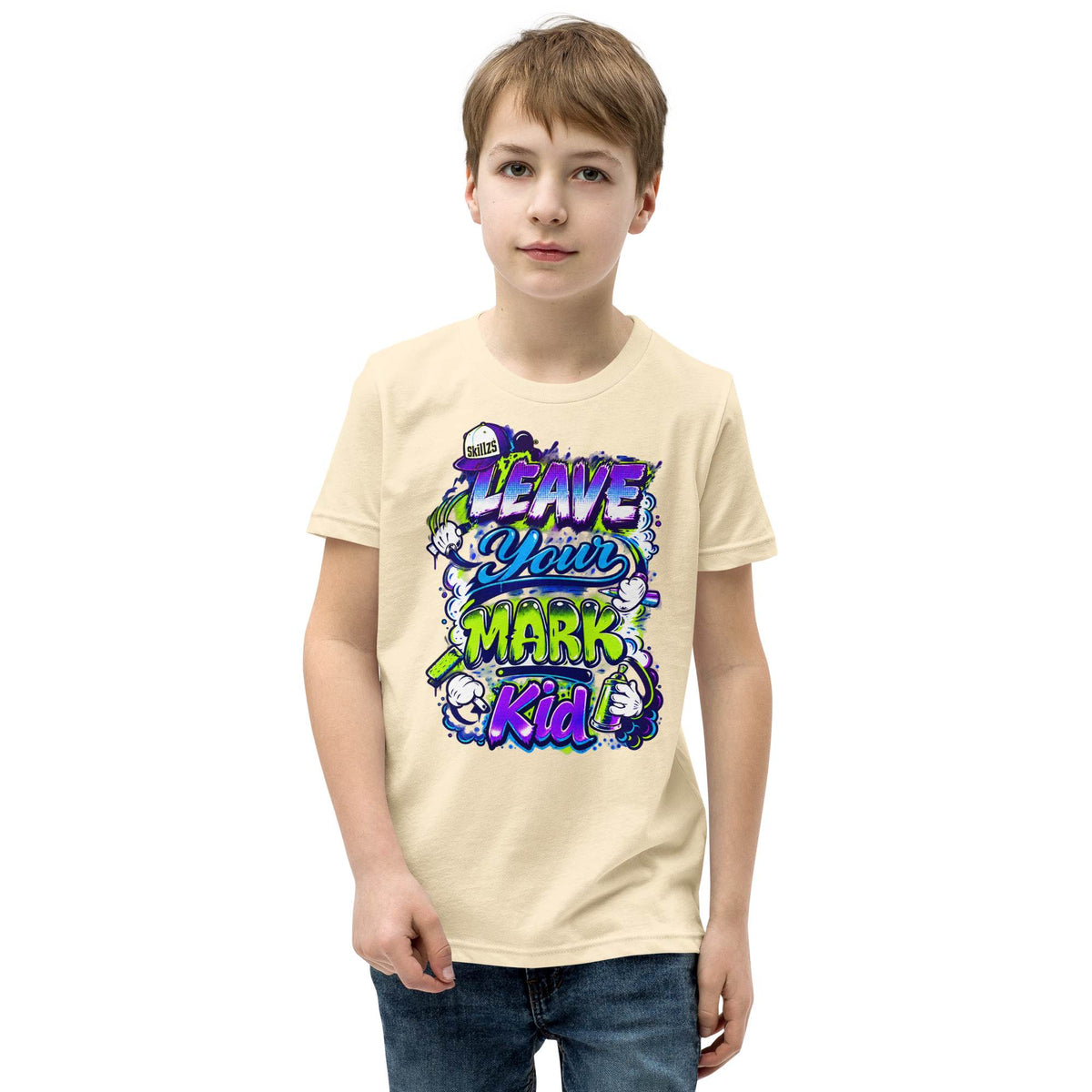 A young boy wearing a light beige graphic t-shirt with a colorful 'Leave Your Mark Kid' design, paired with blue jeans, against a plain white background.