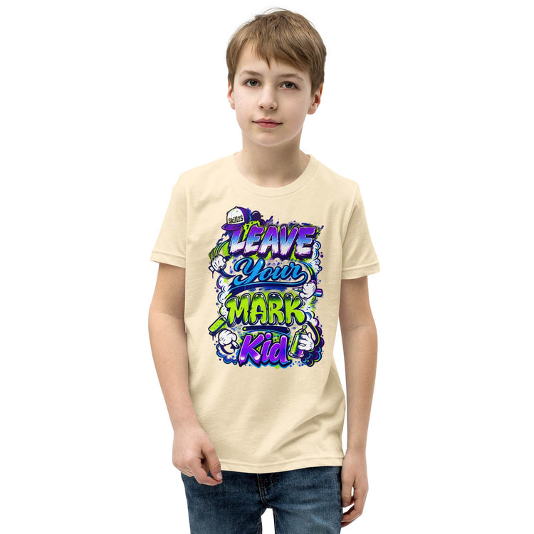 A young boy wearing a light beige graphic t-shirt with a colorful 'Leave Your Mark Kid' design, paired with blue jeans, against a plain white background.