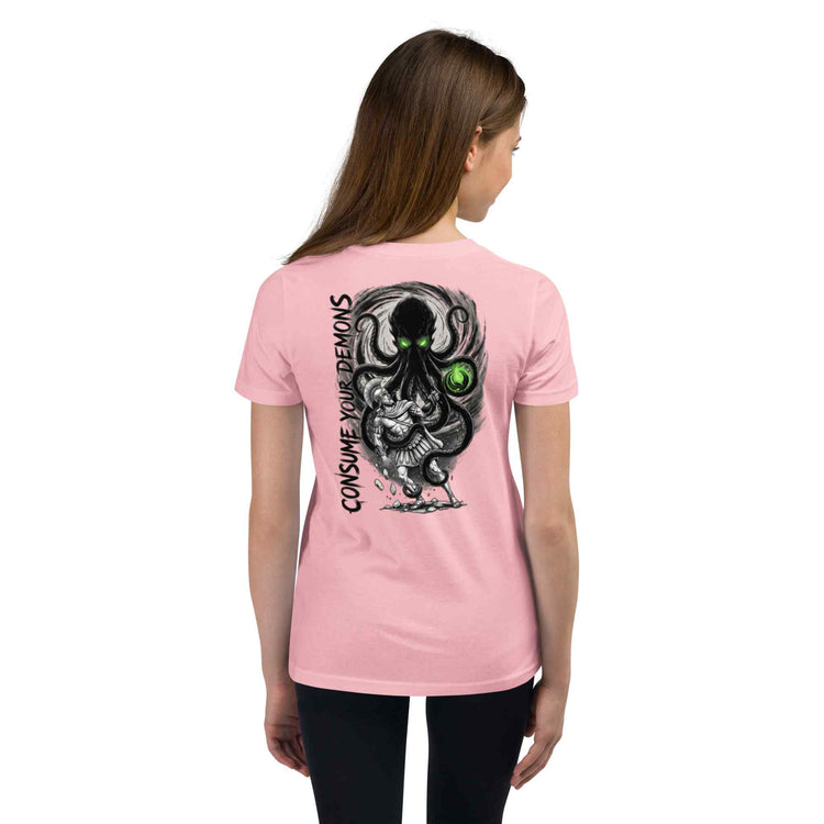 A woman viewed from the back wearing a light pink short-sleeve T-shirt with a large black and gray graphic print on the back featuring a monster and green glowing eyes, plus vertical text reading 'CONSUME YOUR DEMONS'.