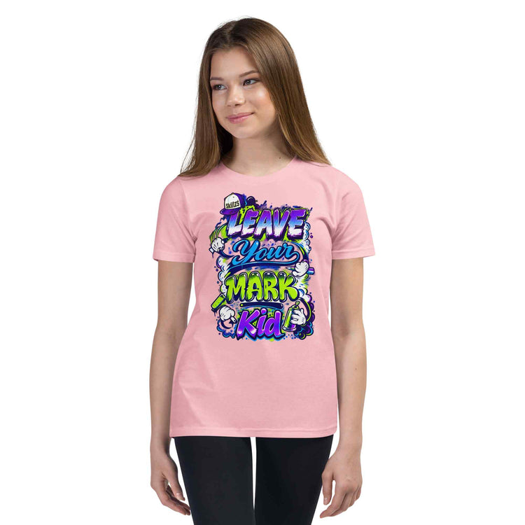 A young woman modeling a light pink t-shirt with a colorful graphic that reads 'LEAVE YOUR MARK Kid' in stylized purple, blue, and green lettering with cartoon characters, set against a plain white background.