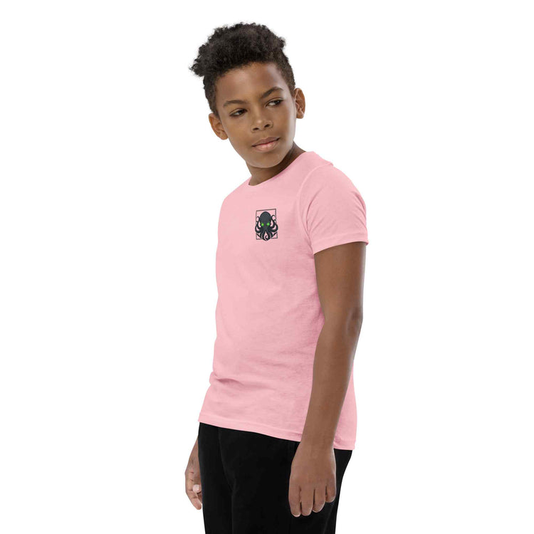 A child model wearing a light pink short-sleeve t-shirt with a small black kraken graphic on the left chest, paired with black pants, set against a plain white background.