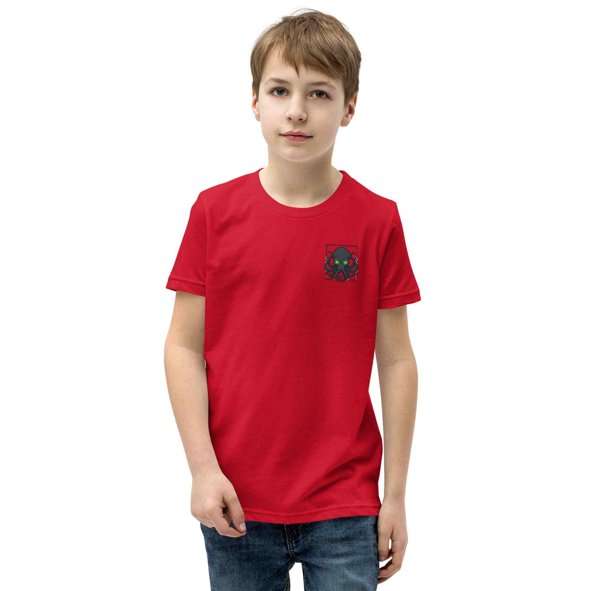 A young boy wearing a bright red short-sleeve t-shirt with a small Kraken graphic on the left chest, paired with blue jeans, photographed against a solid white background.