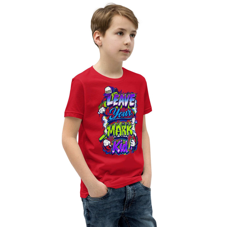 A young boy wearing a bright red graphic t-shirt with a colorful cartoon design that reads 'LEAVE YOUR MARK Kid!'. He is also wearing blue jeans, standing against a plain white background.